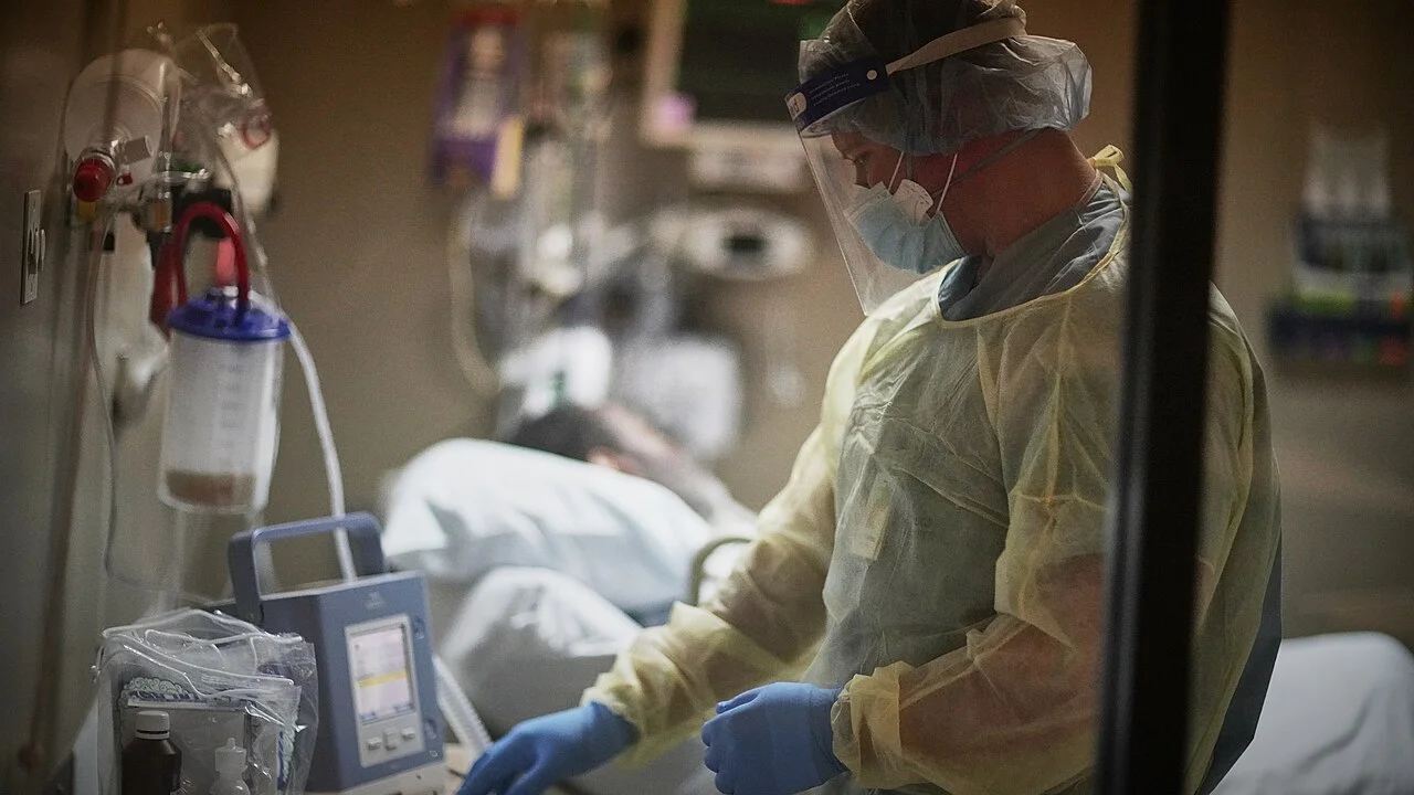 Intensivzimmer im Halbdunkel: Eine Pflegekraft in Schutzkittel, Maske und Visier steht am Bett eines beatmeten Patienten; im Vordergrund ein Gerät mit Display, an der Wand ein Absaugbehälter.