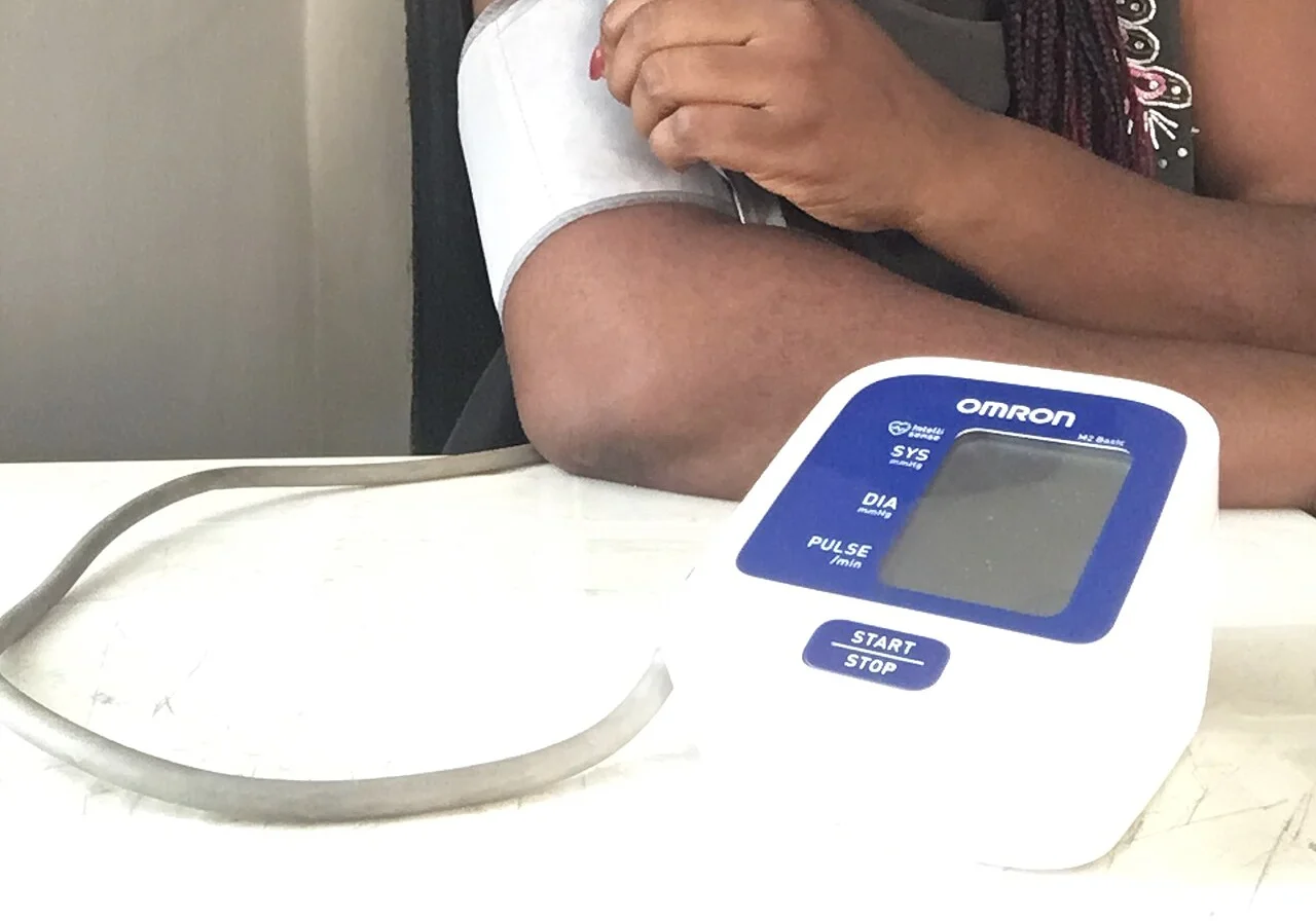 Eine Person legt sich eine Oberarmmanschette an; davor steht ein automatisches Blutdruckmessgerät mit leerem Display und Start-Taste.