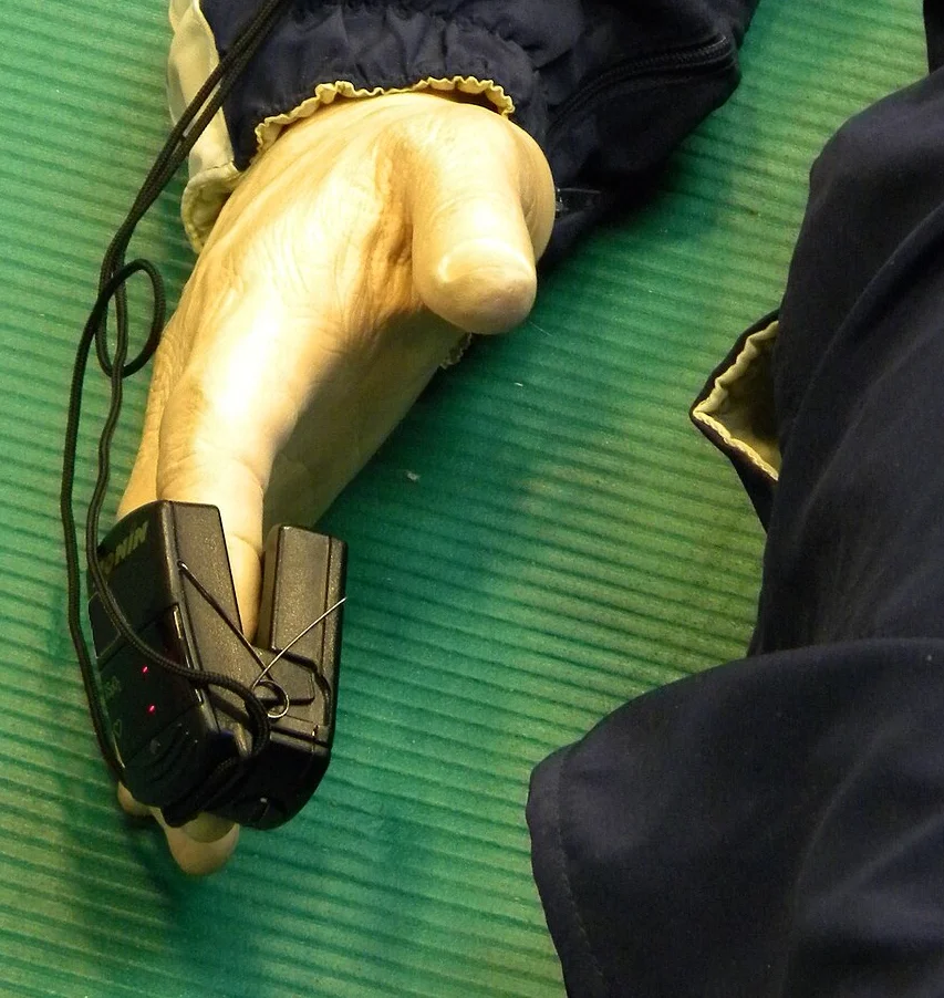 Hand einer Übungspuppe auf einer grünen Matte; an einem Finger sitzt ein schwarzer Fingerclip-Sensor, dessen Kabel am Arm entlang zum Gerät führt.