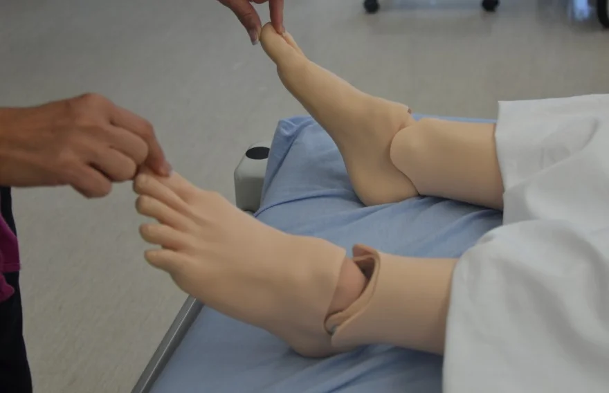 Foto an einer Übungspuppe: Eine Hand drückt kurz auf die Zehenkuppe, um die Rekapillarisierung zu prüfen.