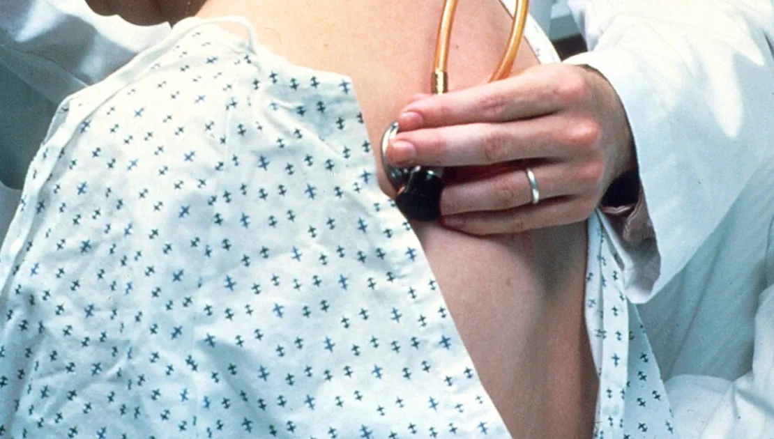 Rückenansicht einer sitzenden Person im hinten geöffneten Patientenhemd; eine Hand hält das Bruststück eines Stethoskops direkt auf die Haut des oberen Rückens.