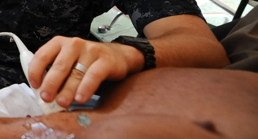 Echtes Foto einer Untersuchung von außen: Eine Hand hält einen Ultraschallschallkopf mit Gel auf der nackten Brustwand einer liegenden Person; das Kabel führt zum Gerät.