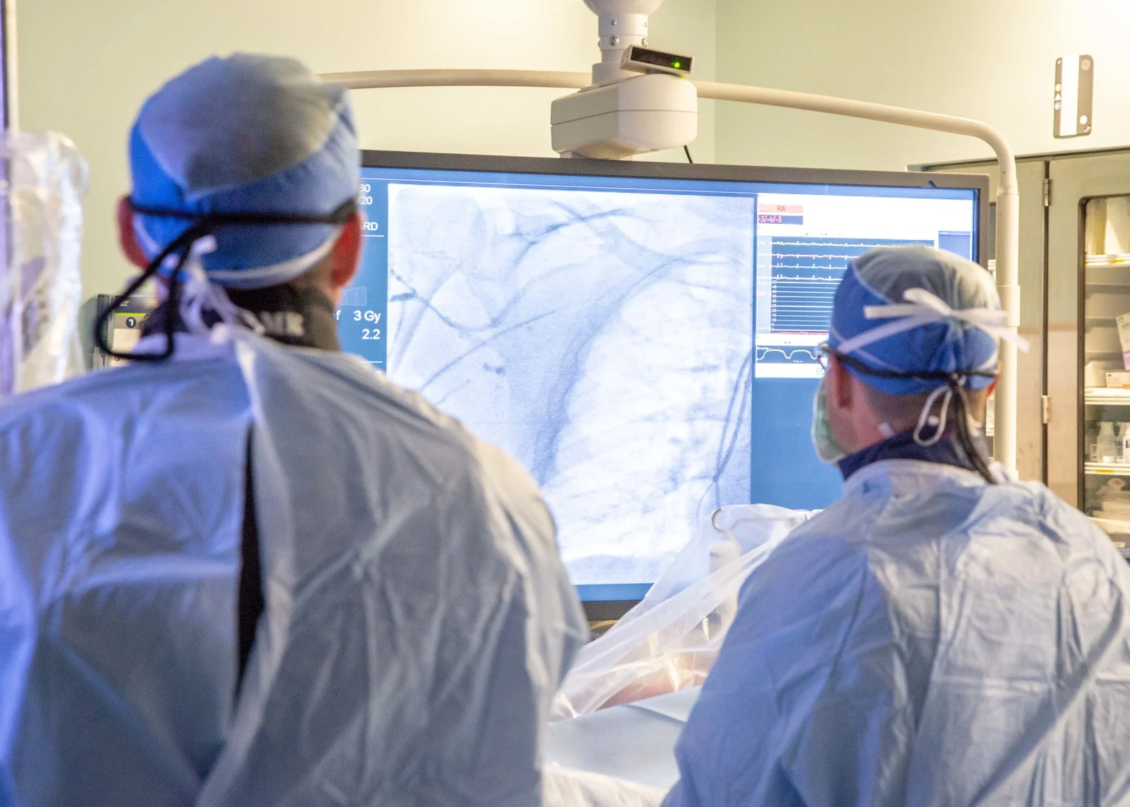 Herzkatheterlabor: zwei Personen in steriler Kleidung mit Haube blicken auf einen großen Monitor mit Durchleuchtungsbild kontrastierter Gefäße und Kreislaufkurven