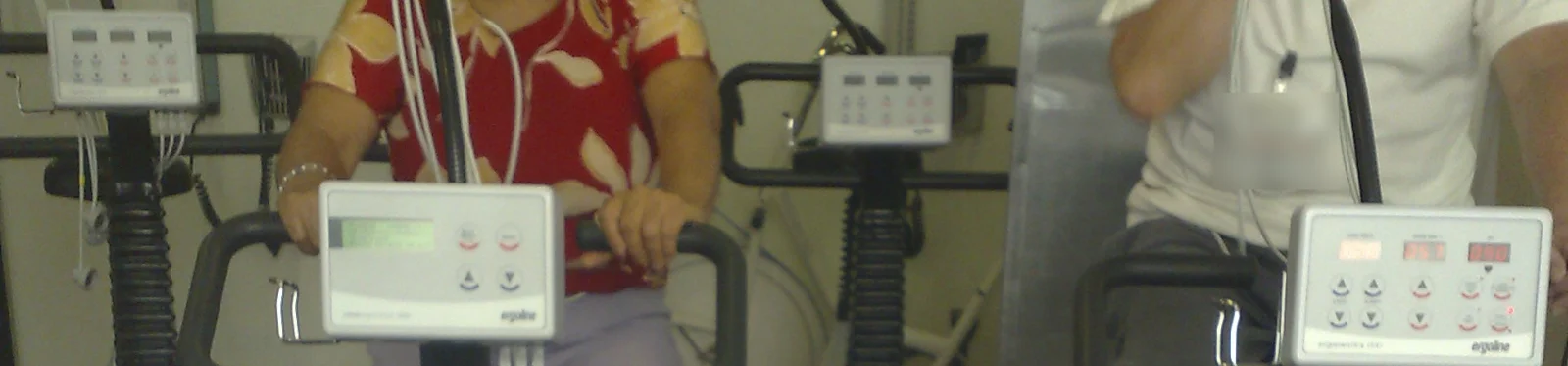 Patientin und Patient beim Ergometertraining im Rahmen einer kardiologischen Rehabilitation, mit EKG-Kabeln am Oberkörper und Bedienkonsolen der Ergometer