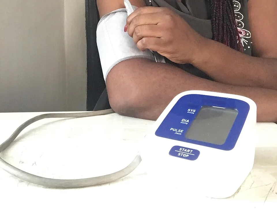 Eine sitzende Person legt sich eine Manschette am freien Oberarm an; der Arm ruht auf dem Tisch, davor steht ein automatisches Oberarmgerät mit Anzeigefeldern für systolischen und diastolischen Wert sowie Puls.