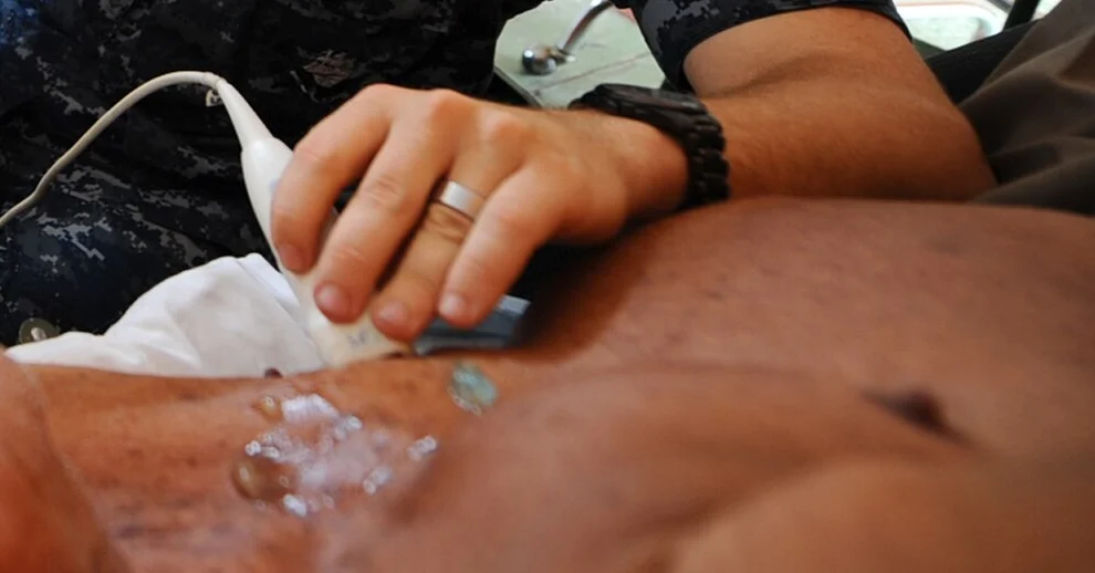Hand mit Ultraschallschallkopf auf der Brustwand eines liegenden Patienten; auf der Haut ist Ultraschallgel zu sehen