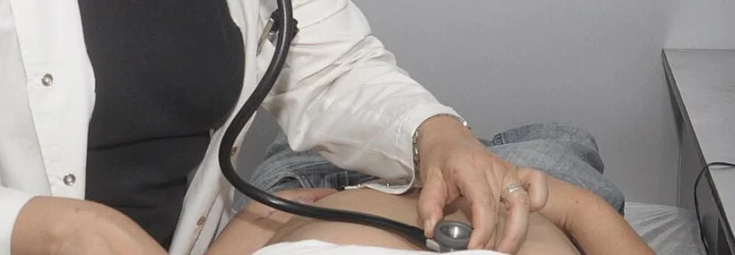 Ausschnitt: Die Hand einer Ärztin hält das Bruststück eines Stethoskops direkt auf die Haut am Brustkorb eines liegenden Patienten; das T-Shirt ist hochgeschoben