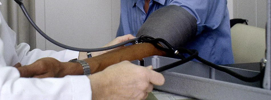 Foto-Ausschnitt: Blutdruckmessung bei einem sitzenden Mann; die Manschette liegt am Oberarm, das Stethoskop führt in die Ellenbeuge, der Arm liegt entspannt auf einer Unterlage.