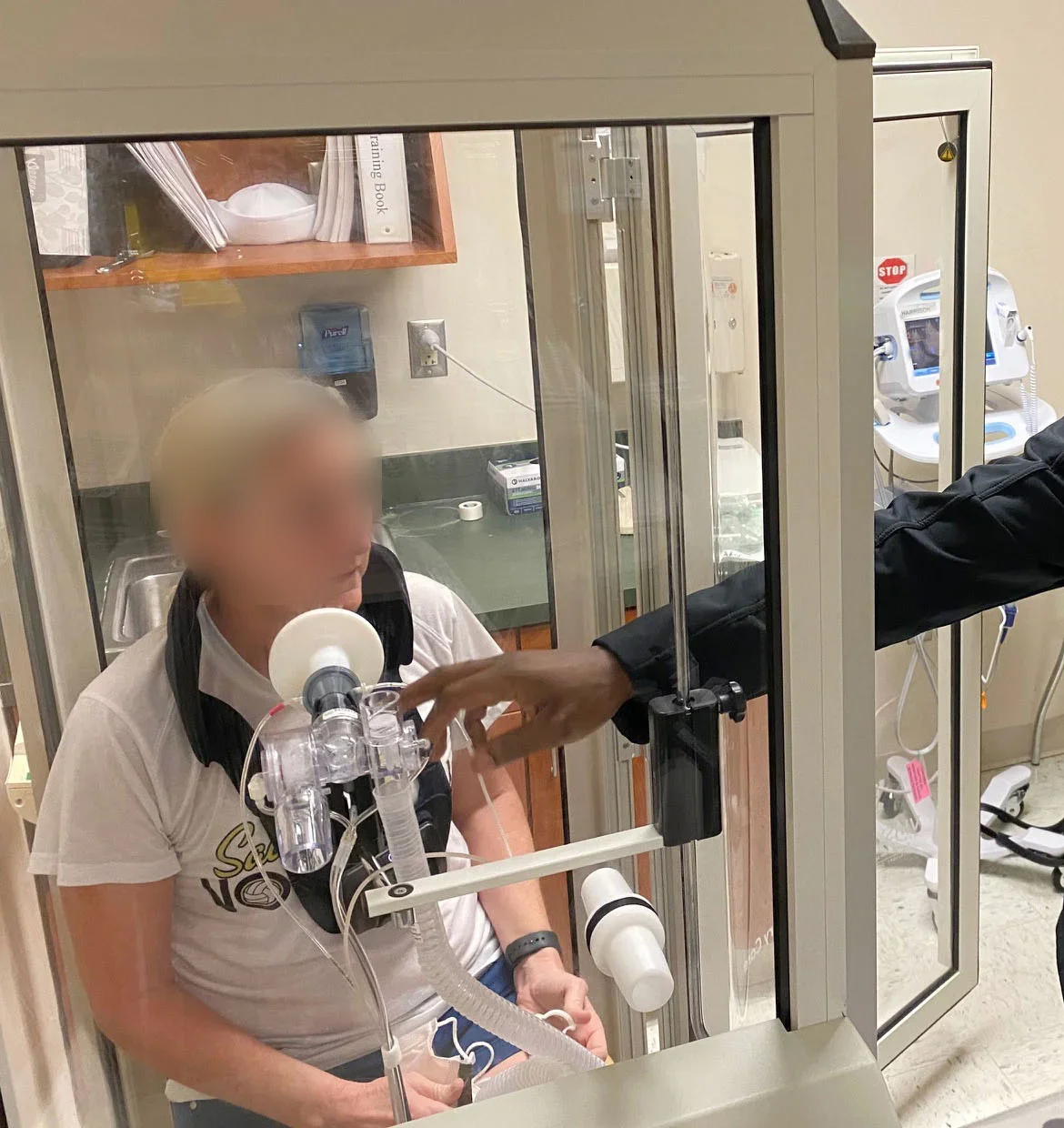 Foto: Eine Patientin sitzt in einer durchsichtigen Messkabine, deren Tür zur Vorbereitung noch offen ist, und hat das Mundstück mit Bakterienfilter und Verschlussventil vor dem Mund; eine Fachkraft greift von außen an die Halterung des Mundstücks.