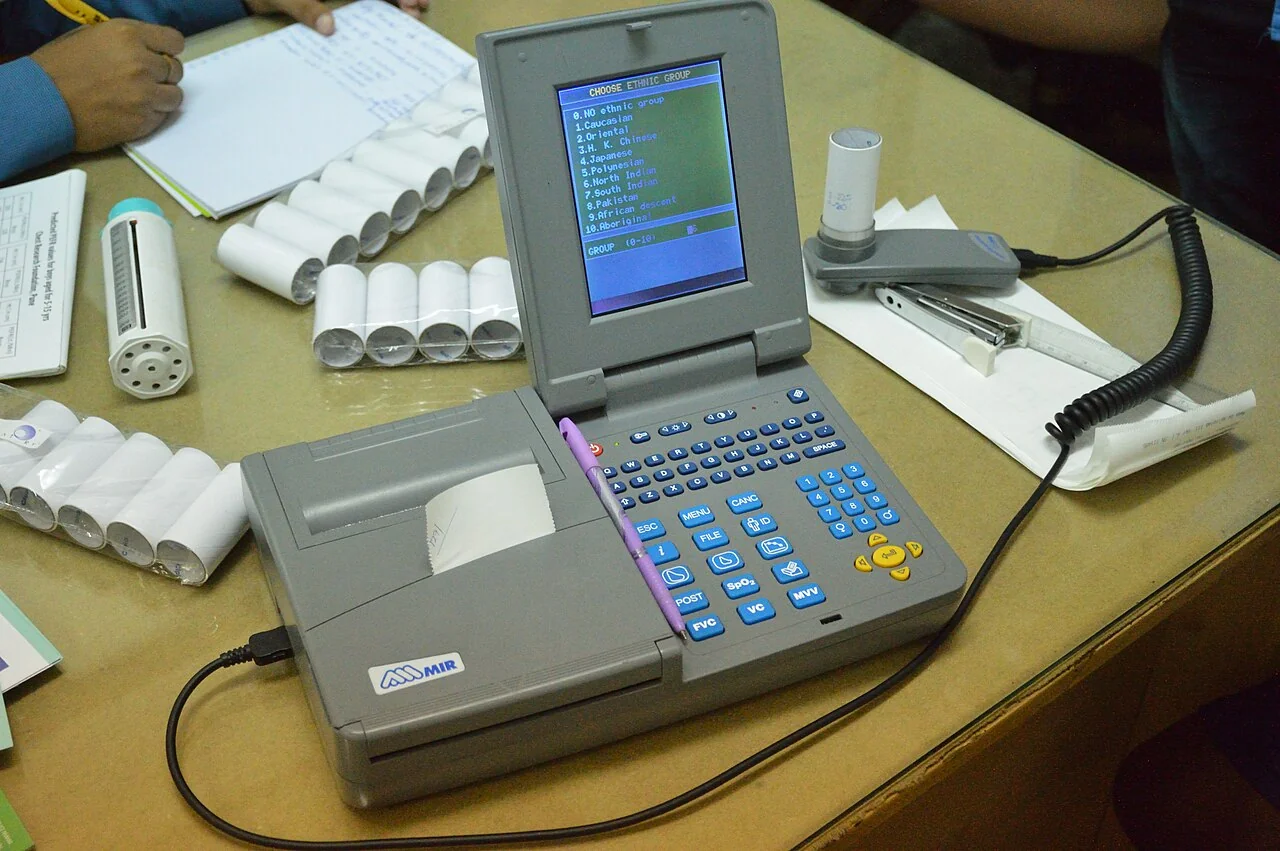 Tragbares Spirometer mit Tastatur, Bildschirm und eingebautem Drucker; daneben ein Durchflusssensor mit Kabel und mehrere Einweg-Papiermundstücke.