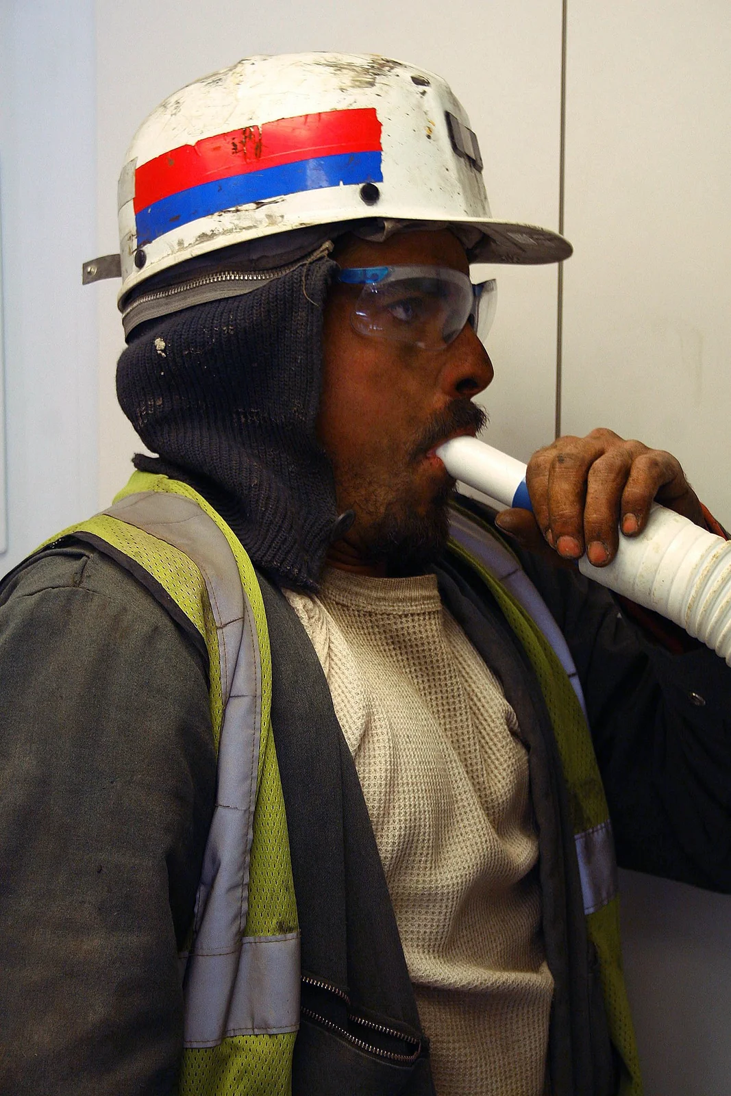 Ein Mann mit Schutzhelm und Schutzbrille atmet über ein weißes Einmalmundstück in den Sensor eines tragbaren Spirometers.
