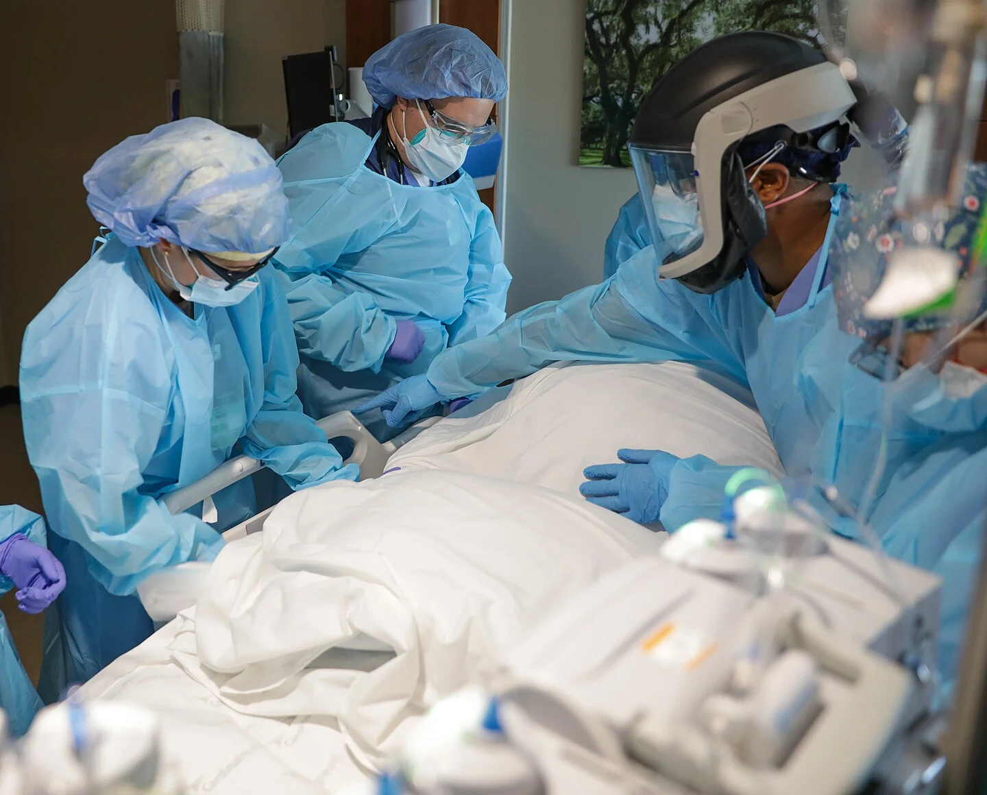 Intensivteam in Schutzkleidung steht von beiden Seiten am Bett und dreht einen in Laken gehüllten Patienten gemeinsam; im Vordergrund Infusionstechnik und Leitungen.
