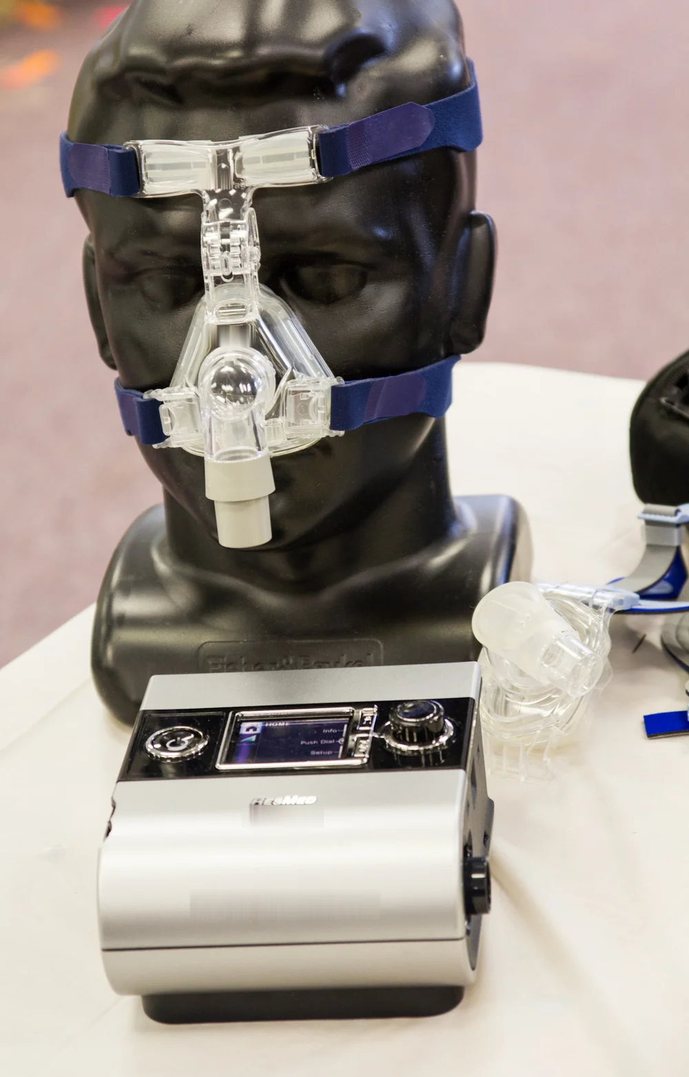 Heim-CPAP-Gerät auf einem Tisch, dahinter ein Puppenkopf mit aufgesetzter Nasenmaske und Kopfbändern; daneben liegt eine weitere Maske.