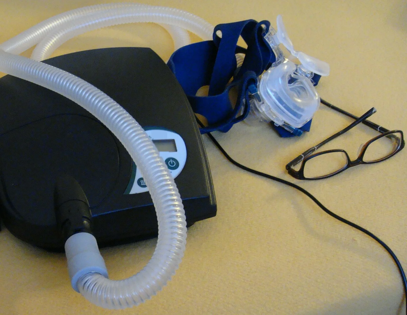 CPAP-Gerät für zu Hause mit Faltenschlauch und Nasenmaske mit Kopfband; eine Brille daneben dient als Größenvergleich.