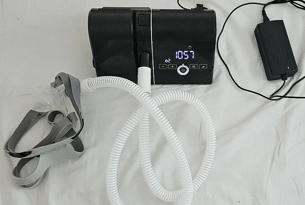 Häusliches PAP-Gerät mit aufgestecktem Befeuchter, weißem Atemschlauch, transparenter Maske mit grauem Kopfband und separatem Netzteil auf einem Bettlaken.