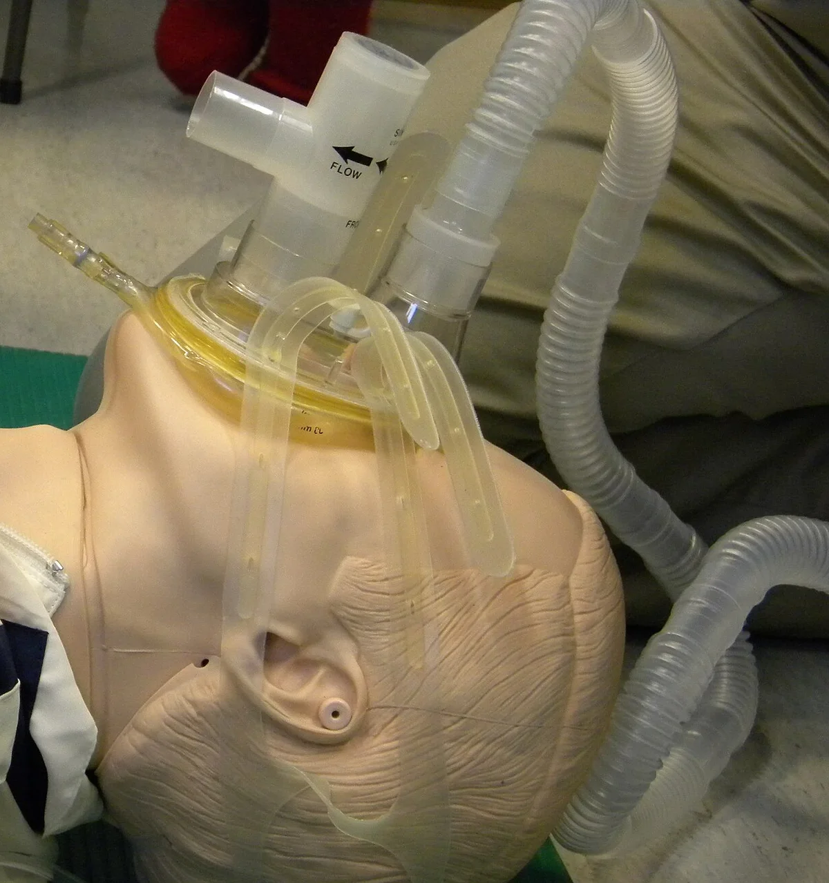 Übungspuppe in Rückenlage mit dicht sitzender Mund-Nasen-Maske, die mit mehreren Gummibändern am Kopf fixiert ist; auf der Maske ein Ventilaufsatz, seitlich Schläuche.