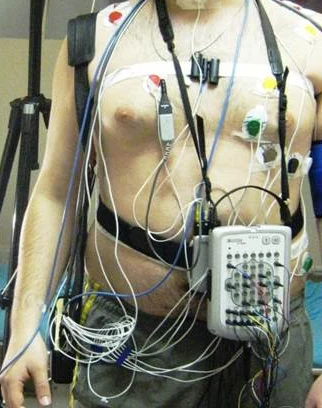 Oberkörper eines Mannes mit angelegter Polysomnographie-Verkabelung: Brust- und Bauchgurt, Elektroden auf Brust und Schultern, ein Sensor am Hals und ein umgehängtes Aufzeichnungsgerät, in dem alle Kabel zusammenlaufen.