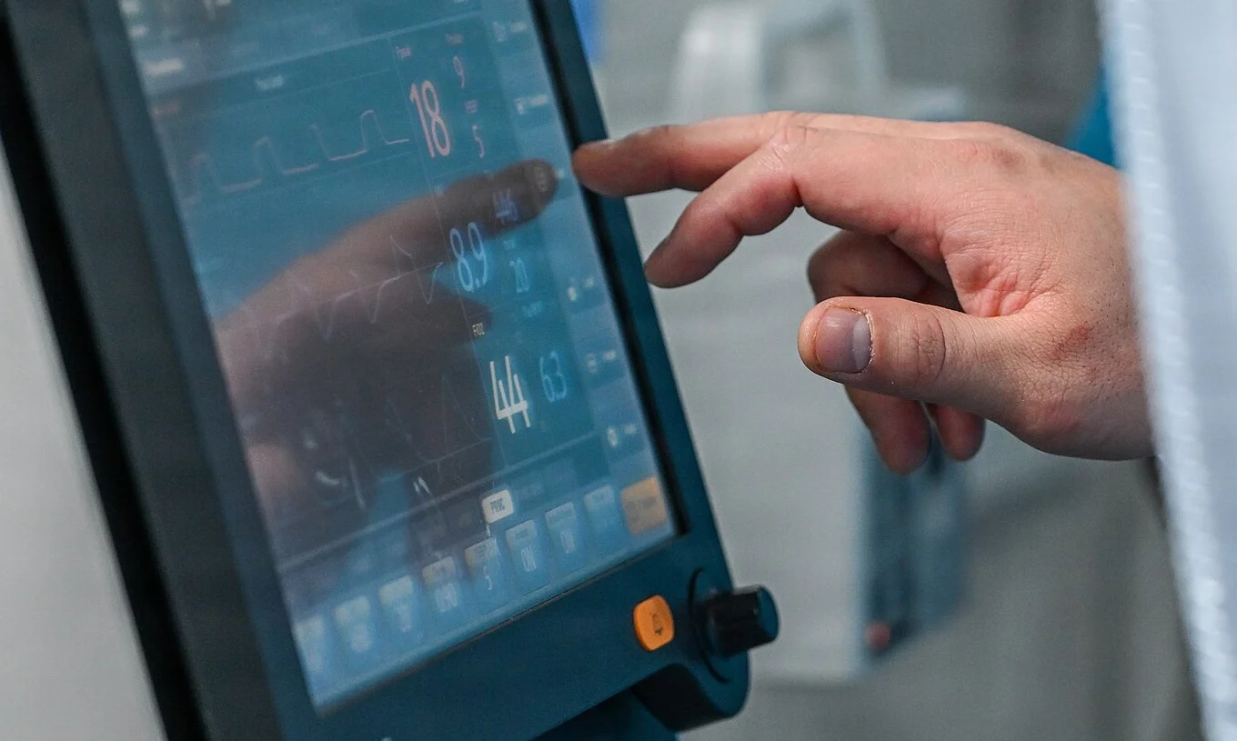 Touchscreen eines Intensivrespirators mit Druckkurve im oberen Feld und großen numerischen Messwerten rechts daneben; eine Hand zeigt auf die Anzeige.