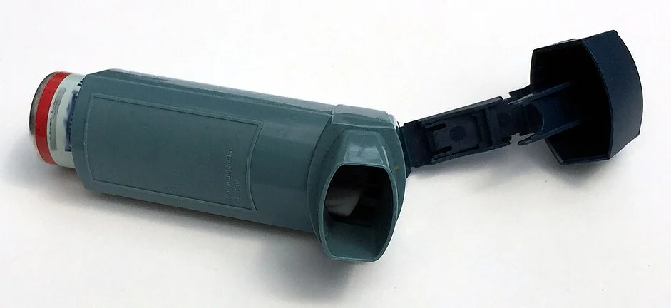 Graublaues Dosieraerosol auf weißem Grund; die Schutzkappe ist abgezogen und hängt am Gehäuse, das Mundstück ist offen, oben steckt der Wirkstoffbehälter mit roter Kappe.