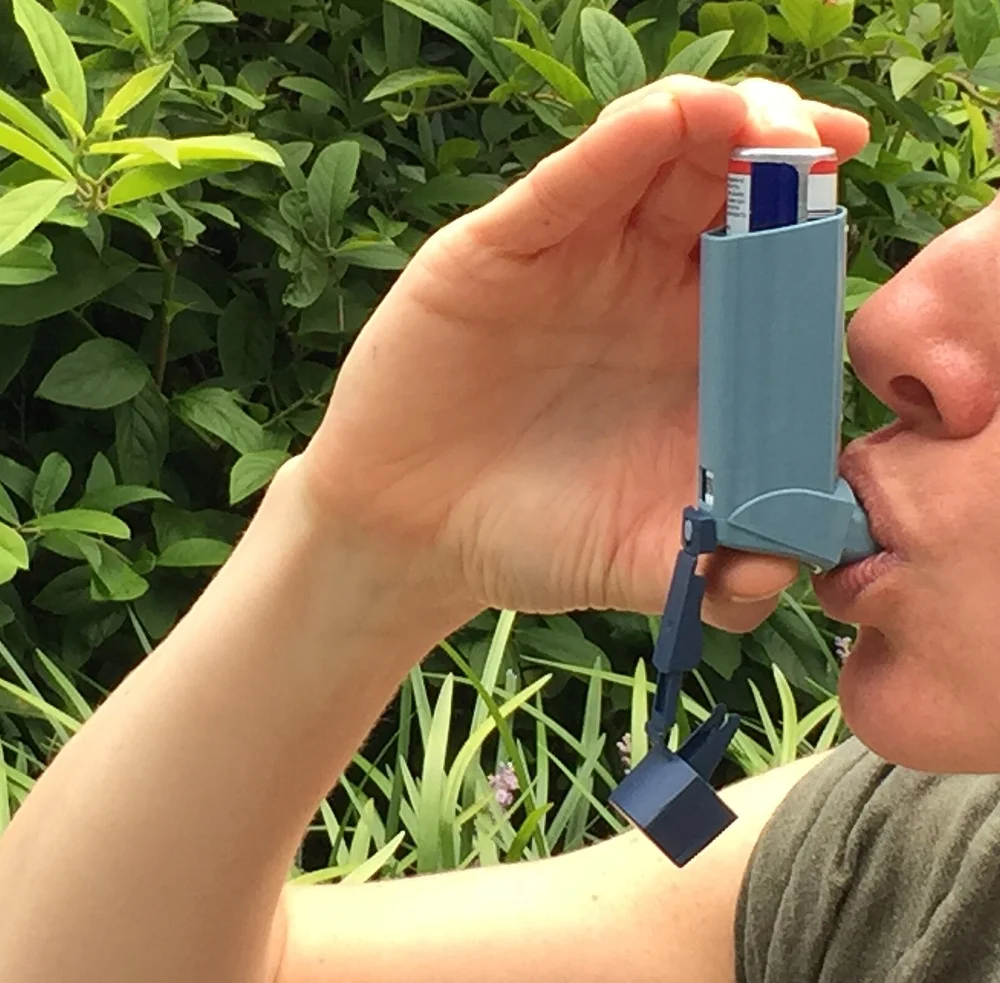 Erwachsene Person hält ein Dosieraerosol senkrecht, die Finger liegen oben auf dem Wirkstoffbehälter, die Lippen umschließen das Mundstück; die abgenommene Schutzkappe hängt an einem Band am Gehäuse.