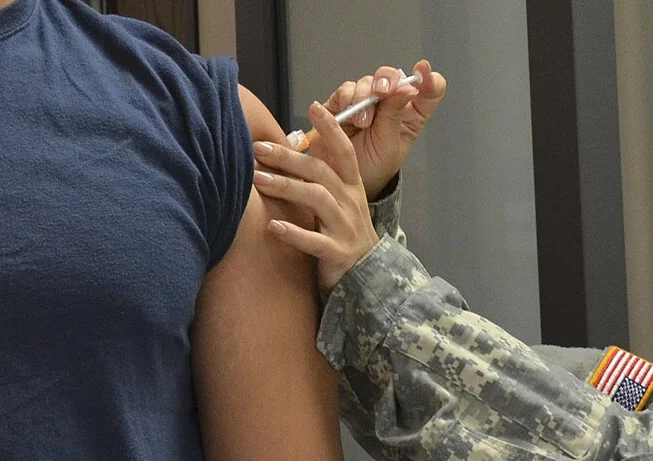 Eine Person in Tarnuniform setzt eine Spritze in den Deltamuskel am Oberarm einer Person mit hochgeschobenem T-Shirt-Ärmel; Gesichter nicht im Bild.