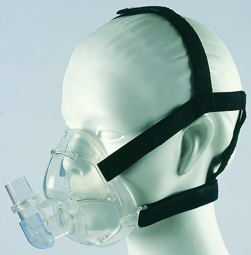 Mund-Nasen-Maske aus transparentem Kunststoff an einem Übungskopf, mit Kopfbändern über Scheitel und Hinterkopf und einem Schlauchanschluss vor dem Mund.