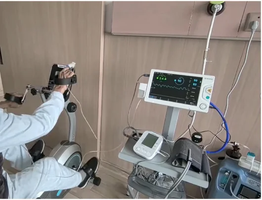 Eine Person sitzt an einem Bewegungstrainer und bewegt Kurbeln mit Armen und Beinen. Daneben stehen ein Patientenmonitor mit Messkurve, ein Blutdruckmessgerät und weitere Überwachungsgeräte.