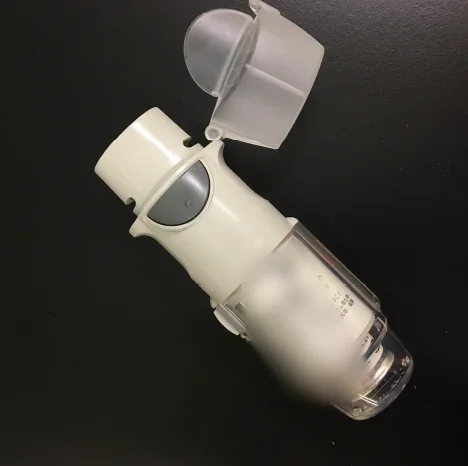 Weißer Respimat-Inhalator mit aufgeklappter durchsichtiger Schutzkappe vor dunklem Hintergrund. Oben das Mundstück, seitlich die graue Auslösetaste, unten das durchsichtige Unterteil mit der eingesetzten Kartusche.