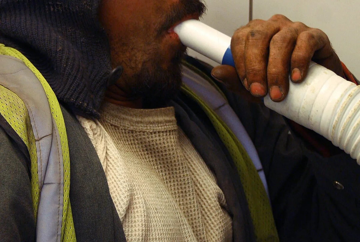 Ein Mann umschließt das weiße Mundstück eines Spirometers fest mit den Lippen und hält den Schlauch mit der Hand; Oberkörper in Arbeitskleidung, Augenpartie nicht im Bild.