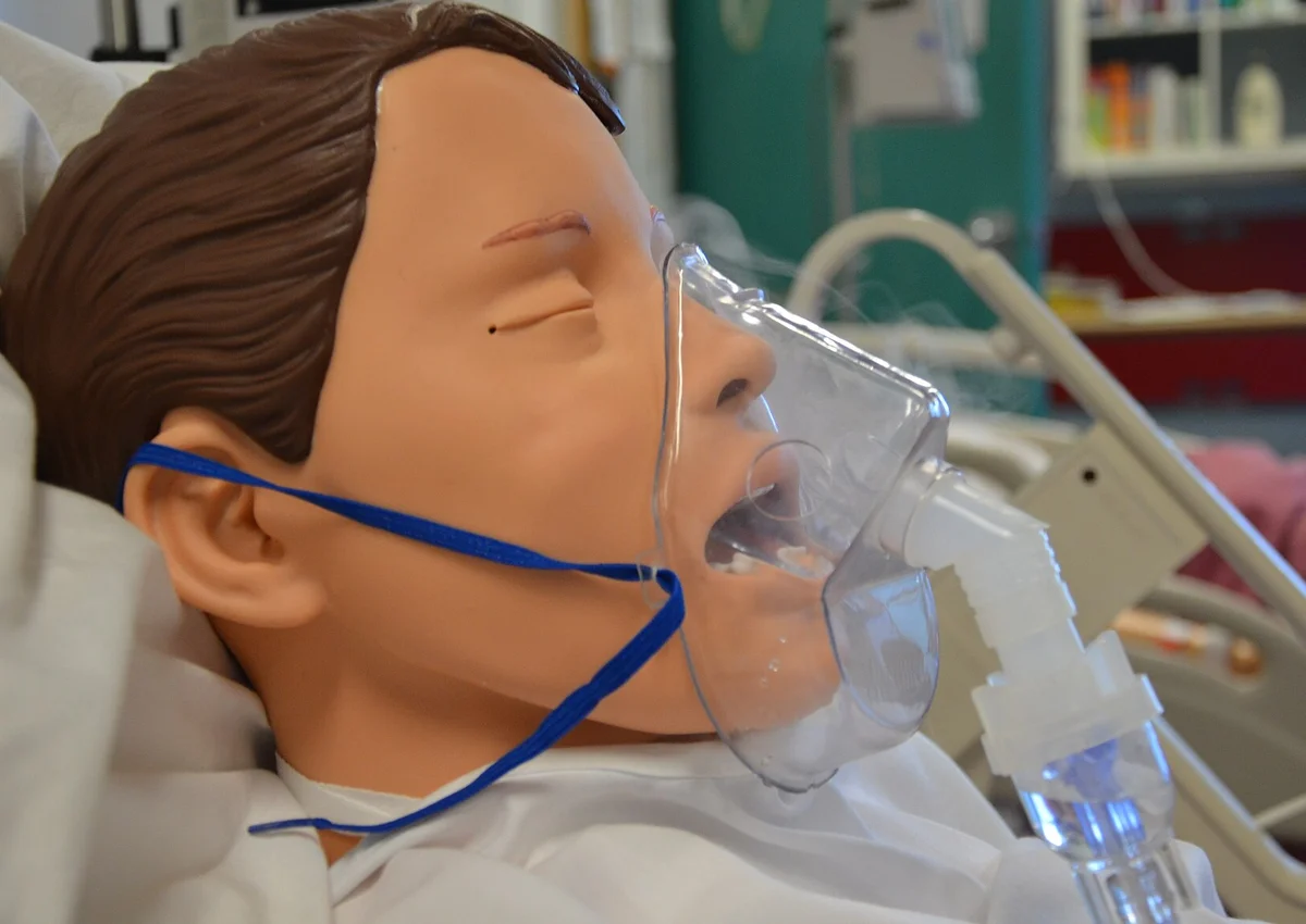 Übungspuppe im Krankenbett mit durchsichtiger Gesichtsmaske über Mund und Nase, die ein Gummiband am Kopf hält; unterhalb der Maske hängt die Verneblerkammer.