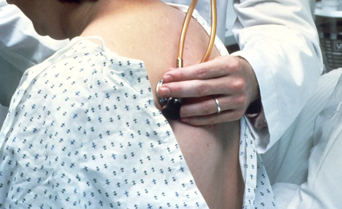 Auskultation am Rücken: Eine Person im Patientenhemd sitzt, die untersuchende Hand setzt die Membran des Stethoskops auf den oberen Rücken.