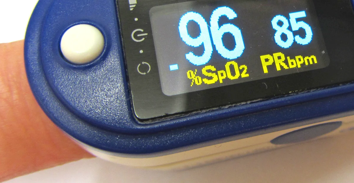 Fingerpulsoxymeter am Finger mit Anzeige der Sauerstoffsättigung von 96 % und einer Pulsfrequenz von 85 pro Minute.