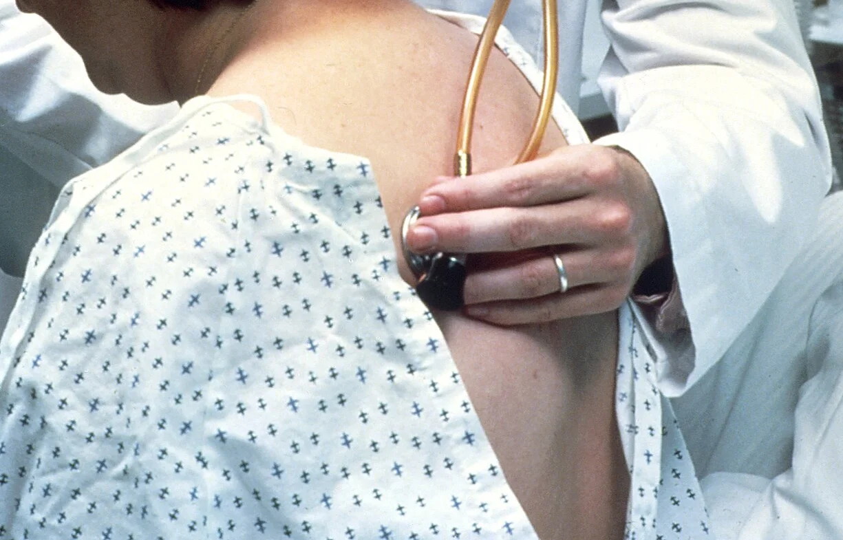 Ärztliche Hand setzt die Membran eines Stethoskops am oberen Rücken einer sitzenden Person im Patientenhemd auf.