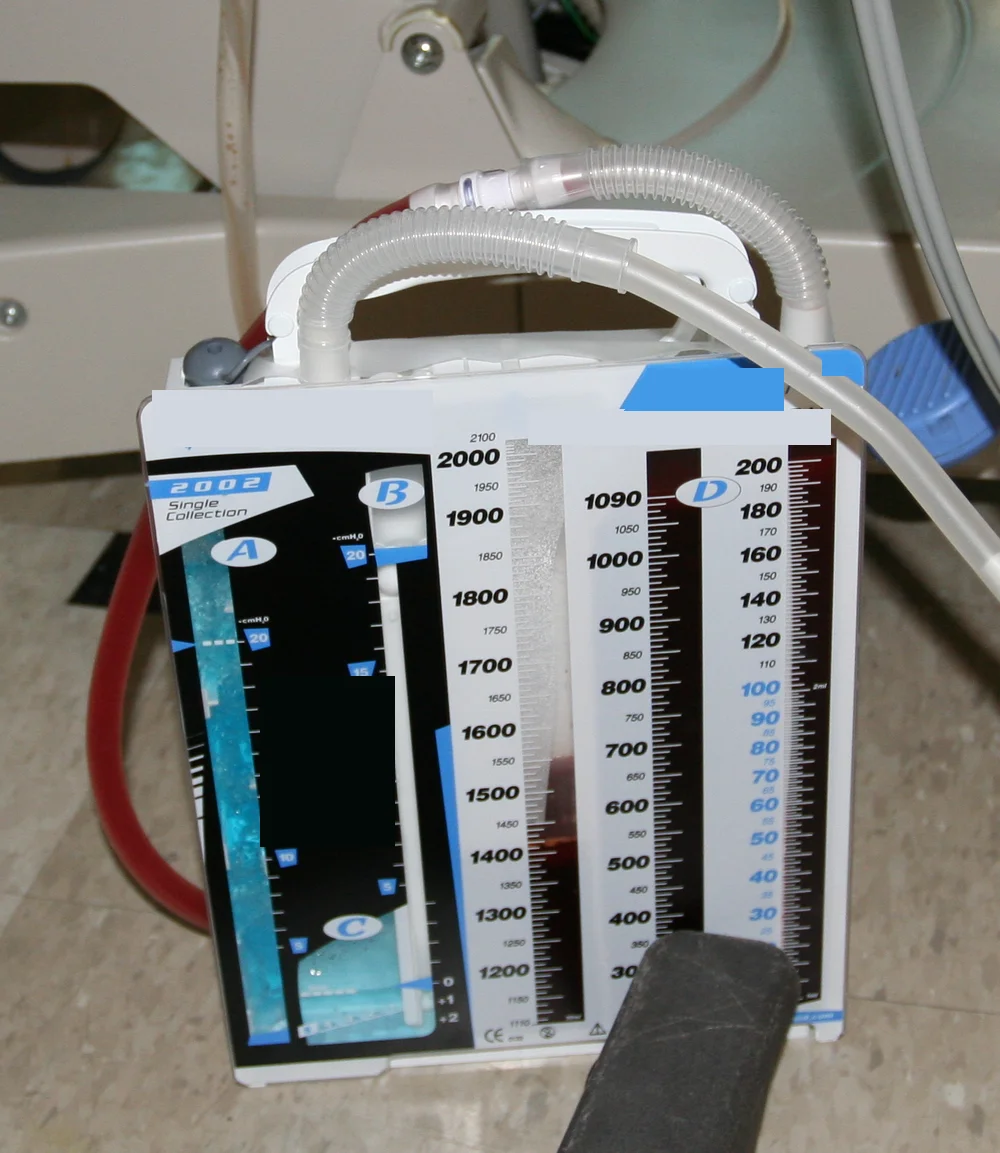 Thoraxdrainage-Sammelsystem mit Wasserschloss steht aufrecht auf dem Boden neben dem Krankenbett; oben führen Schläuche zum Gerät, links verläuft ein dunkelroter Schlauch, vorn sind Kammern mit Skalen zu sehen.