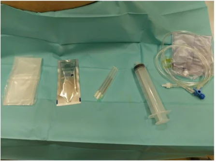 Auf einem grünen sterilen Tuch liegen nebeneinander eine verpackte sterile Schutzhülle, ein kleiner verpackter Beutel, Punktionskanülen, eine große Spritze und ein Schlauchsystem mit Dreiwegehahn und Auffangbeutel.