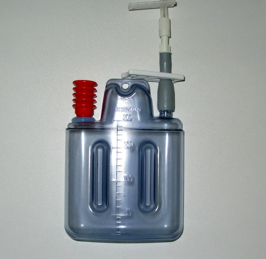 Durchsichtige Wunddrainageflasche mit Milliliterskala bis 200 ml, rotem Faltenbalg und Schlauchanschluss mit Klemme, an einer hellen Wand hängend.