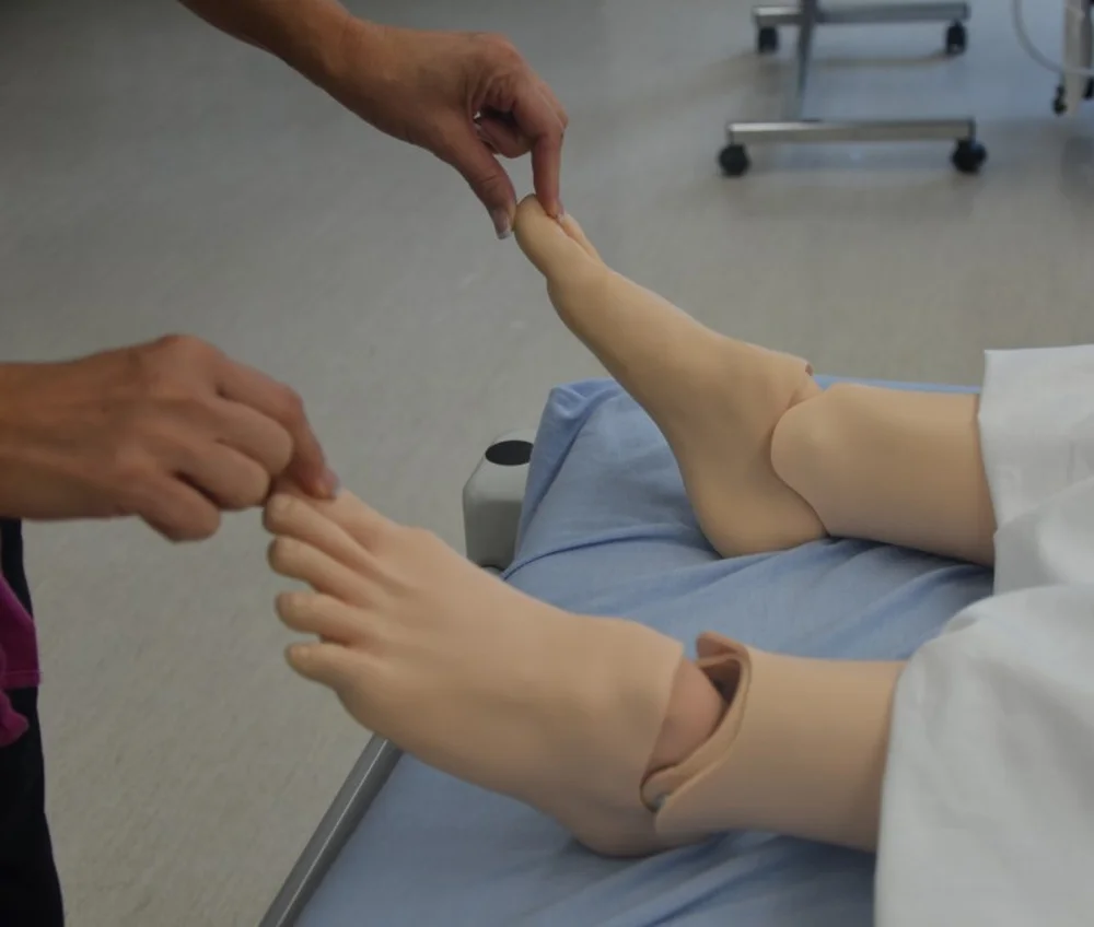 Eine untersuchende Person drückt an einer Übungspuppe mit Daumen und Zeigefinger auf die Großzehe; die andere Hand hält den zweiten Fuß an den Zehen.