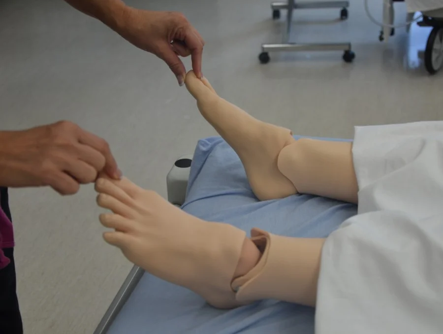 Übungssituation an einer Patientenpuppe auf einer Liege: Eine Untersucherin drückt mit Daumen und Zeigefinger kurz auf eine Großzehe, mit der anderen Hand fasst sie die Zehen des zweiten Fußes.