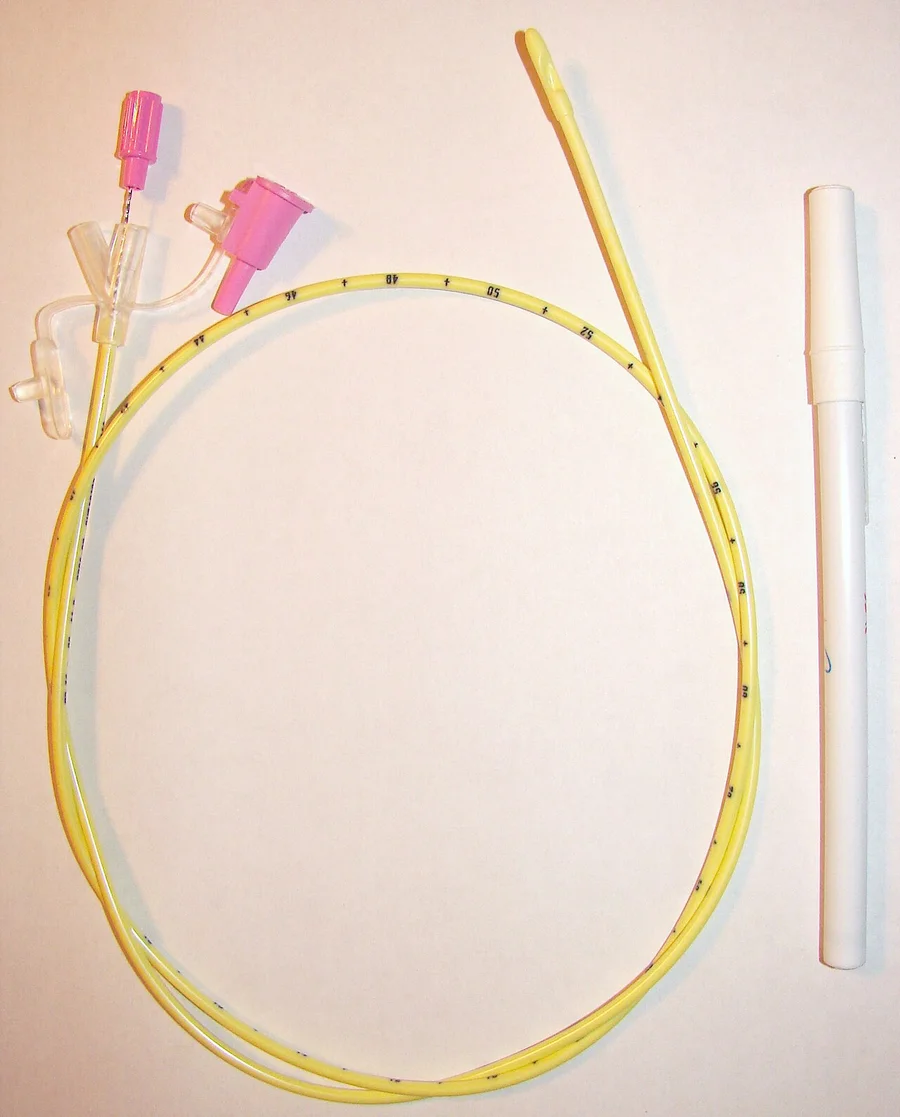 Gelbe, dünne Sonde mit Zentimetermarkierungen, zu einem Ring gelegt, mit pinkfarbenen Anschlüssen und eingelegtem Führungsdraht; daneben eine weiße Schutzhülse.