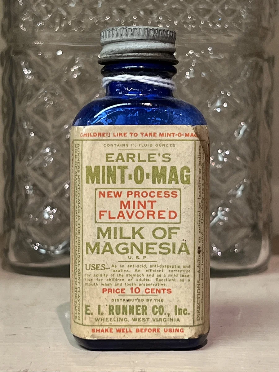 Kleine blaue Glasflasche mit vergilbtem Papieretikett „Milk of Magnesia“ und aufgedruckten Hinweisen zur Anwendung als Abführ- und Säurebindungsmittel.