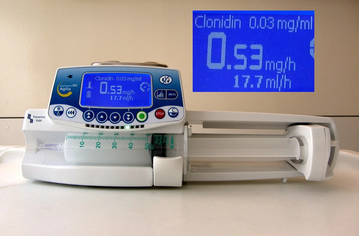 Spritzenpumpe mit eingelegter 50-ml-Spritze; das Display zeigt einen Medikamentennamen mit Konzentration in mg/ml, darunter eine Angabe in mg/h und eine Angabe in ml/h; rechts oben ist das Display vergrößert eingeblendet.