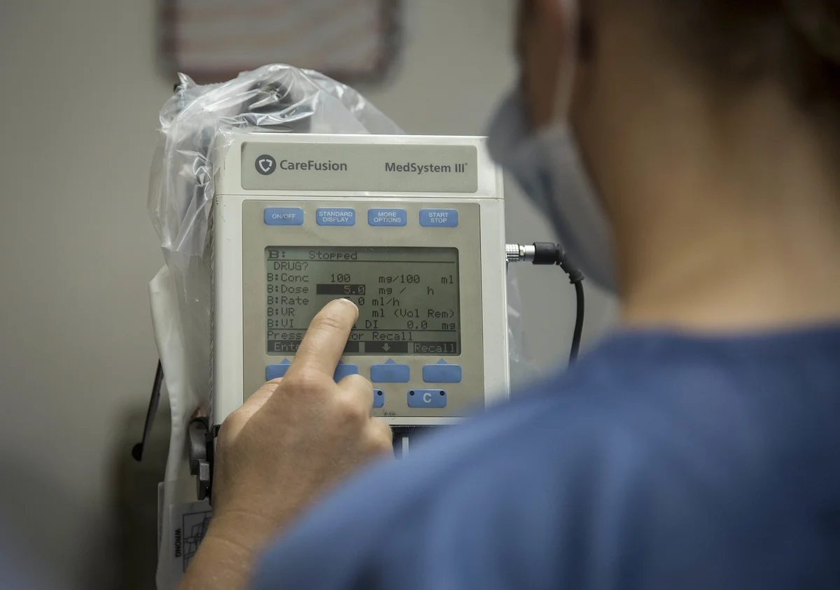 Ein Finger bedient die Tasten einer Infusionspumpe; das Display zeigt untereinander Zeilen für Konzentration in mg/100 ml, Dosis in mg/h und Rate in ml/h.