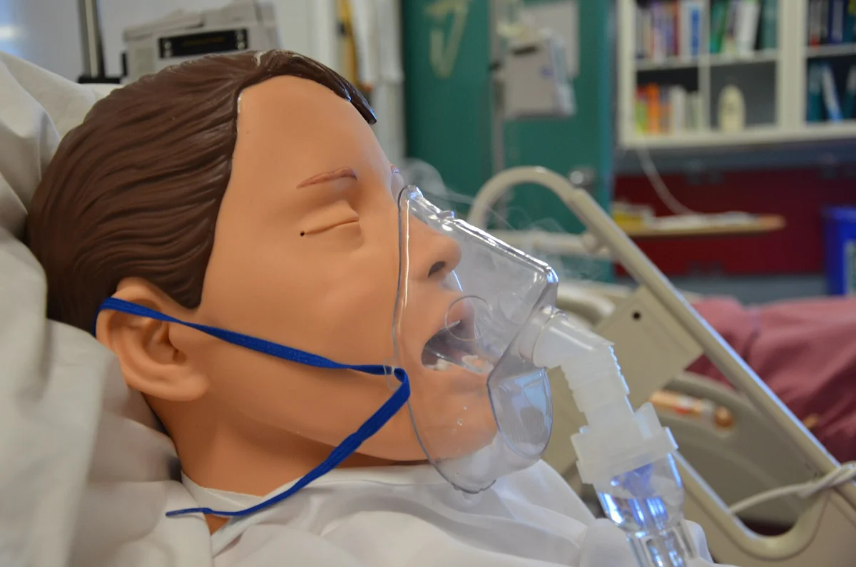 Übungspuppe in einem Klinikbett mit durchsichtiger Gesichtsmaske, die über einen Schlauchansatz mit einem kleinen Medikamentenbehälter unterhalb des Kinns verbunden ist.