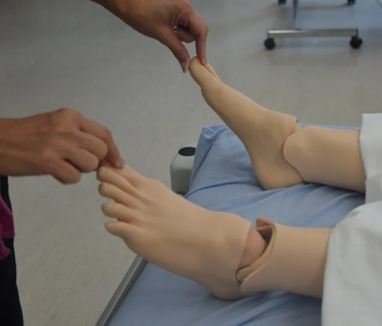 Eine untersuchende Hand drückt kurz auf die Großzehe einer liegenden Übungspuppe, um die Rekapillarisierungszeit zu prüfen; die andere Hand hält die Zehe des zweiten Fußes.