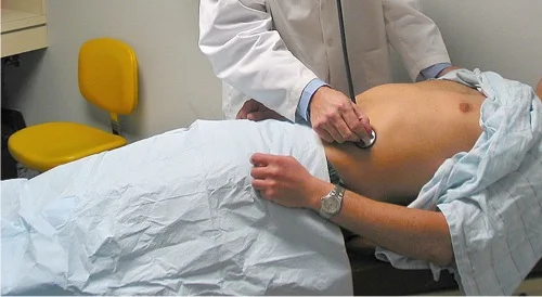 Foto einer Untersuchungssituation: Ein Patient liegt in Rückenlage, Unterkörper und Beine sind mit einem Tuch abgedeckt, nur der Bauch ist frei; die untersuchende Person auskultiert mit dem Stethoskop.