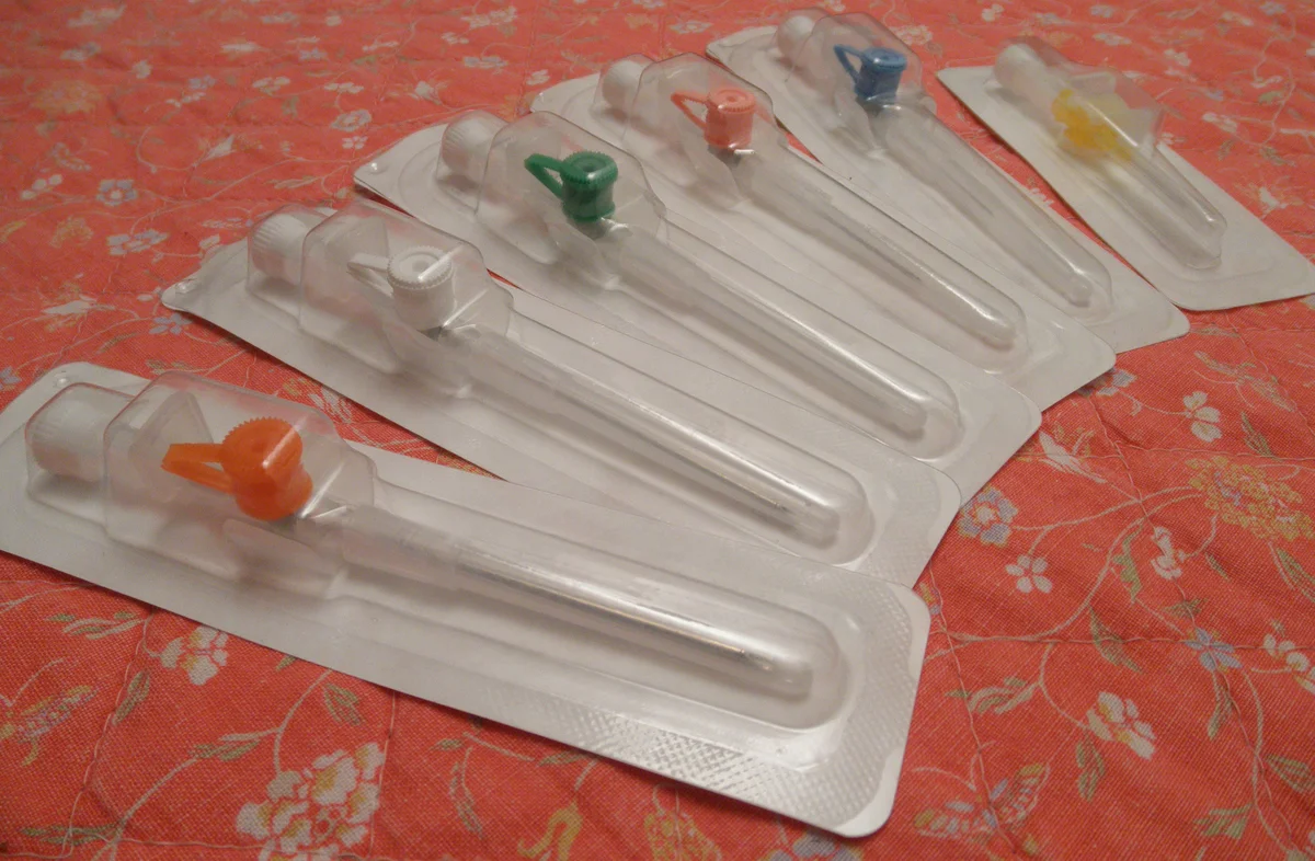 Foto von sechs periphervenösen Verweilkanülen in Blisterverpackungen, nebeneinander gelegt und nach Farbe codiert: orange, weiß, grün, rosa, blau und gelb, von links nach rechts mit abnehmendem Durchmesser.